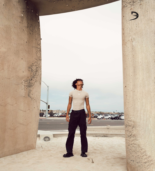Model wearing short sleeve physical tee and black pants staring up at a rock structure standing at the beach.