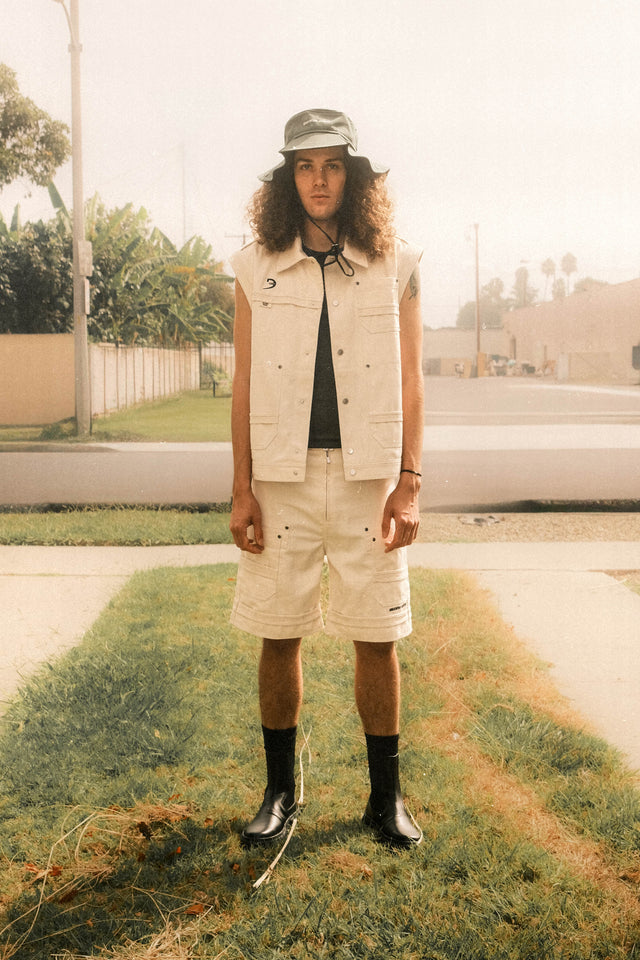 Model wearing evergreen floppy bucket hat, evergreen racer crew tank, bone color vest and bone color cargo shorts standing in front of apartment.
