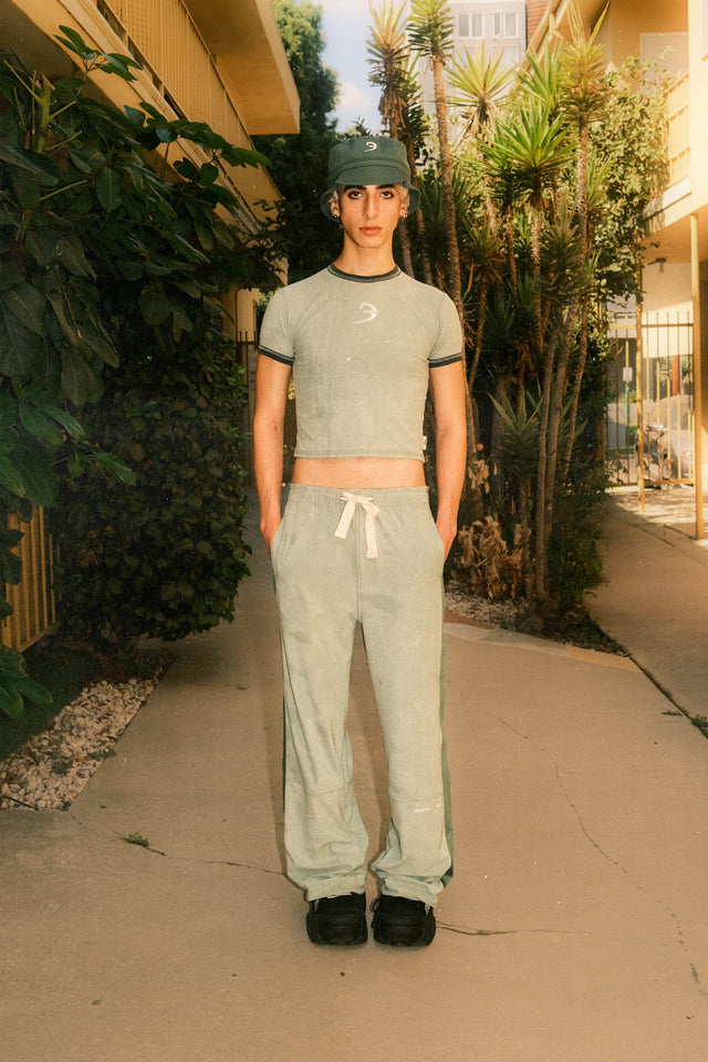 Model wearing evergreen floppy bucket hat with green baby tee and green track pants standing in front of apartment.