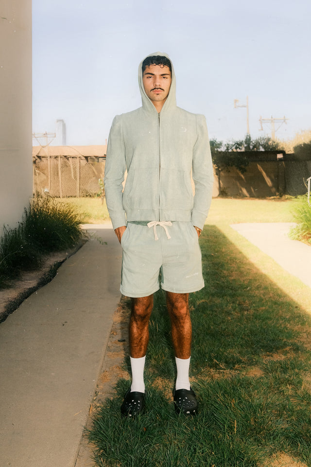 Model wearing hoodie in green with shorts in green standing in front of apartment side yard.