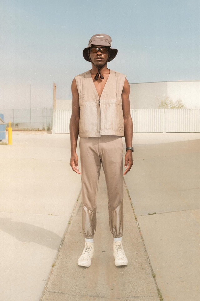 Model wearing floppy bucket hat in graphite with shiny taupe vest and matching pants with elasticized hem standing in front of commercial business driveway.