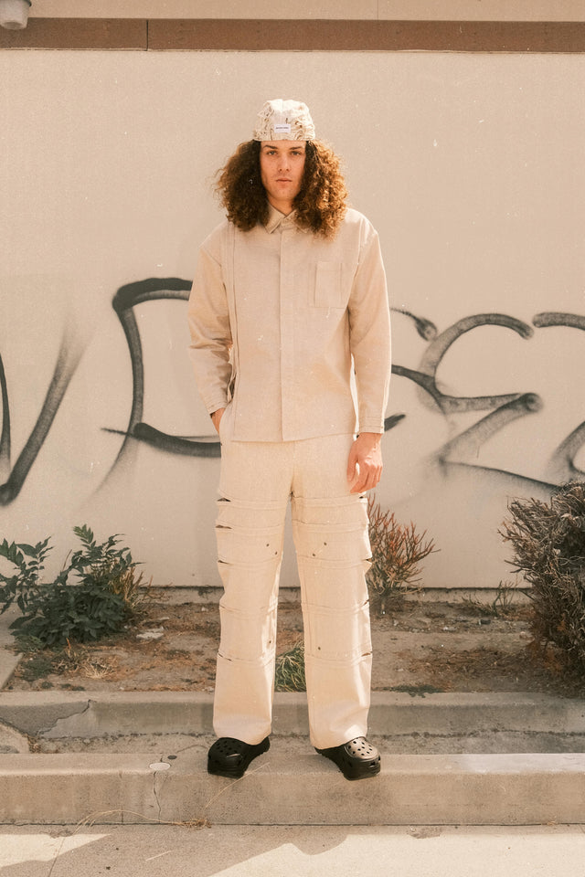 Model wearing bandana hat in brown and cream with ivory button down shirt and linen pants with side cut outs standing in front of graffiti wall.