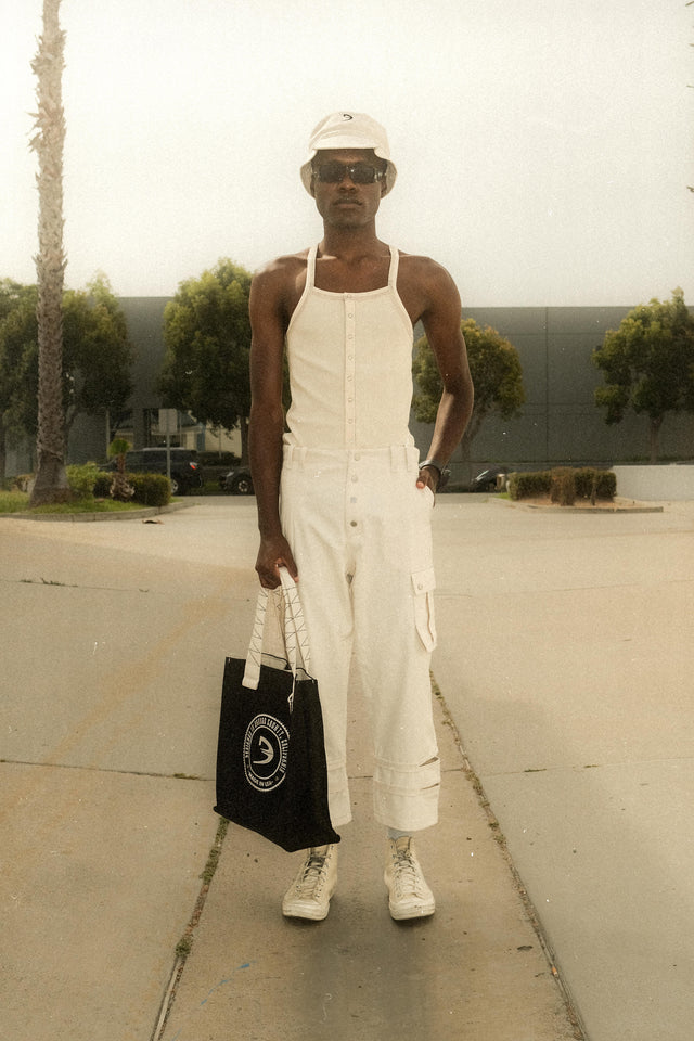 Model wearing tapered bucket hat in bone with ivory color ribbed strappy tank andcream color cargo pants holding black ETX tote bag standing in front of commercial building side driveway.