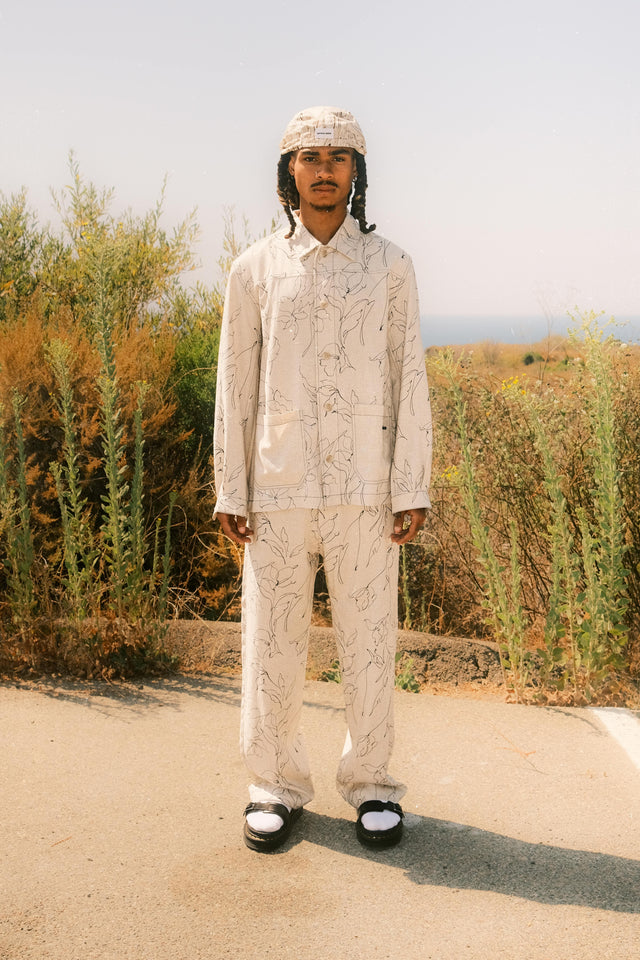 Model wearing bandana hat in brown and cream paired with collared chore jacket in black abstract art print and matching wide leg pants standing in front of flower field.