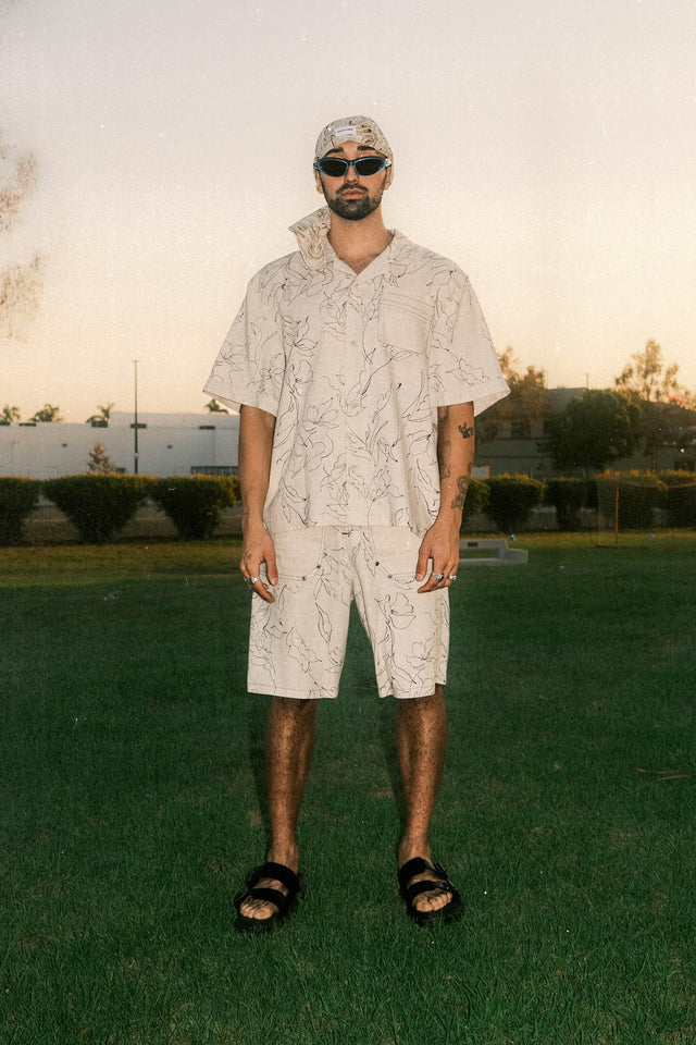 Model wearing bandana hat in black and white colorway paired with resort shirt and cargo shorts in black print against ivory color linen standing in a recreational park.