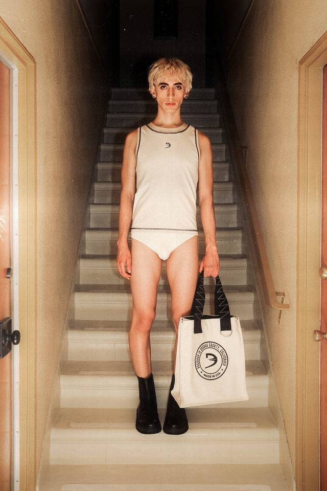 Model wearing racer crew tank in ivory, classic briefs in eggshell and holding ETX tote bag in natural color standing in front of apartment staircase.