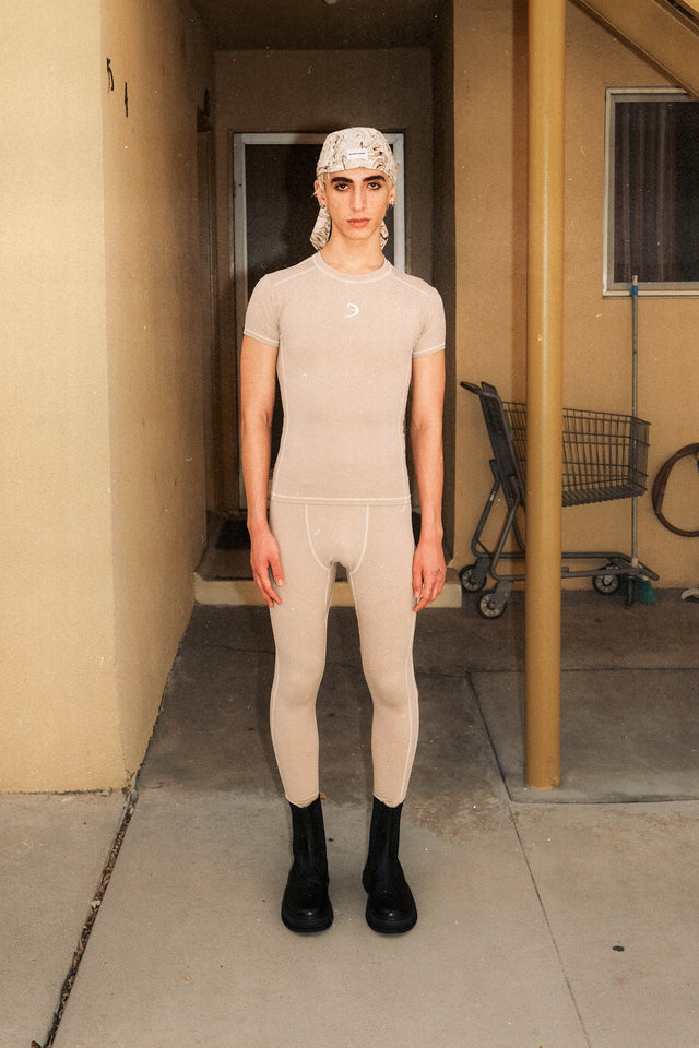 Model in bandana hat in brown and cream, compression shirt in tan and compression tights in tan standing in front of apartment.