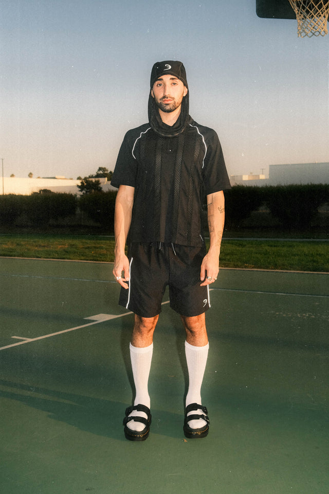 Model wearing black mask as a hat, black mesh top and black mesh shorts standing in a basketball court.