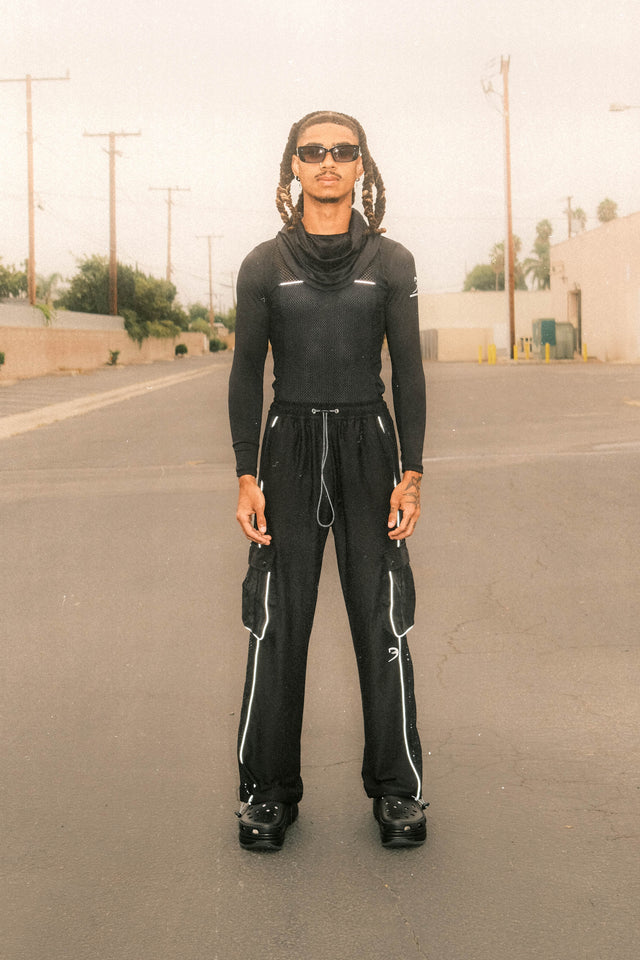 Model wearing black mesh top with black mesh pants standing in front of driveway behind business.