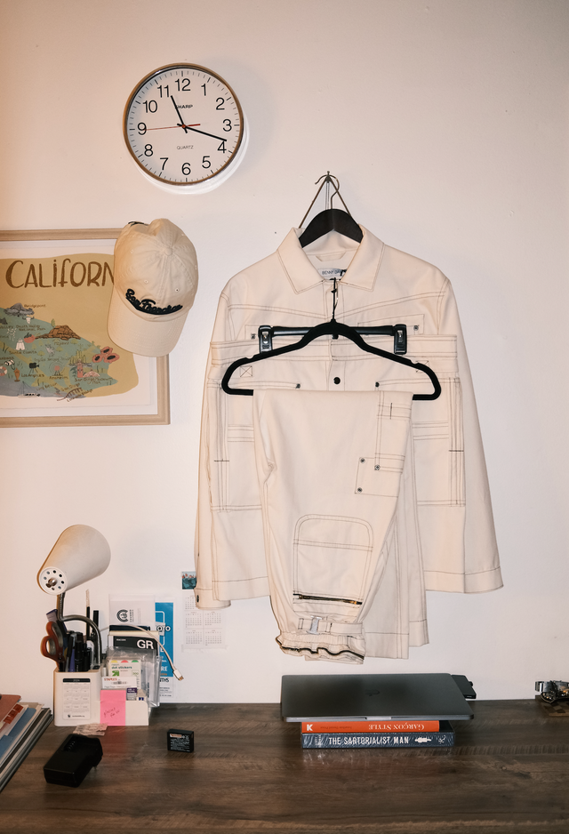 Office view showcasing clock on a wall, California artwork poster with a San Francisco hat hung on the edge, convertible coveralls on hangers.  Table has laptop and some fashion books.